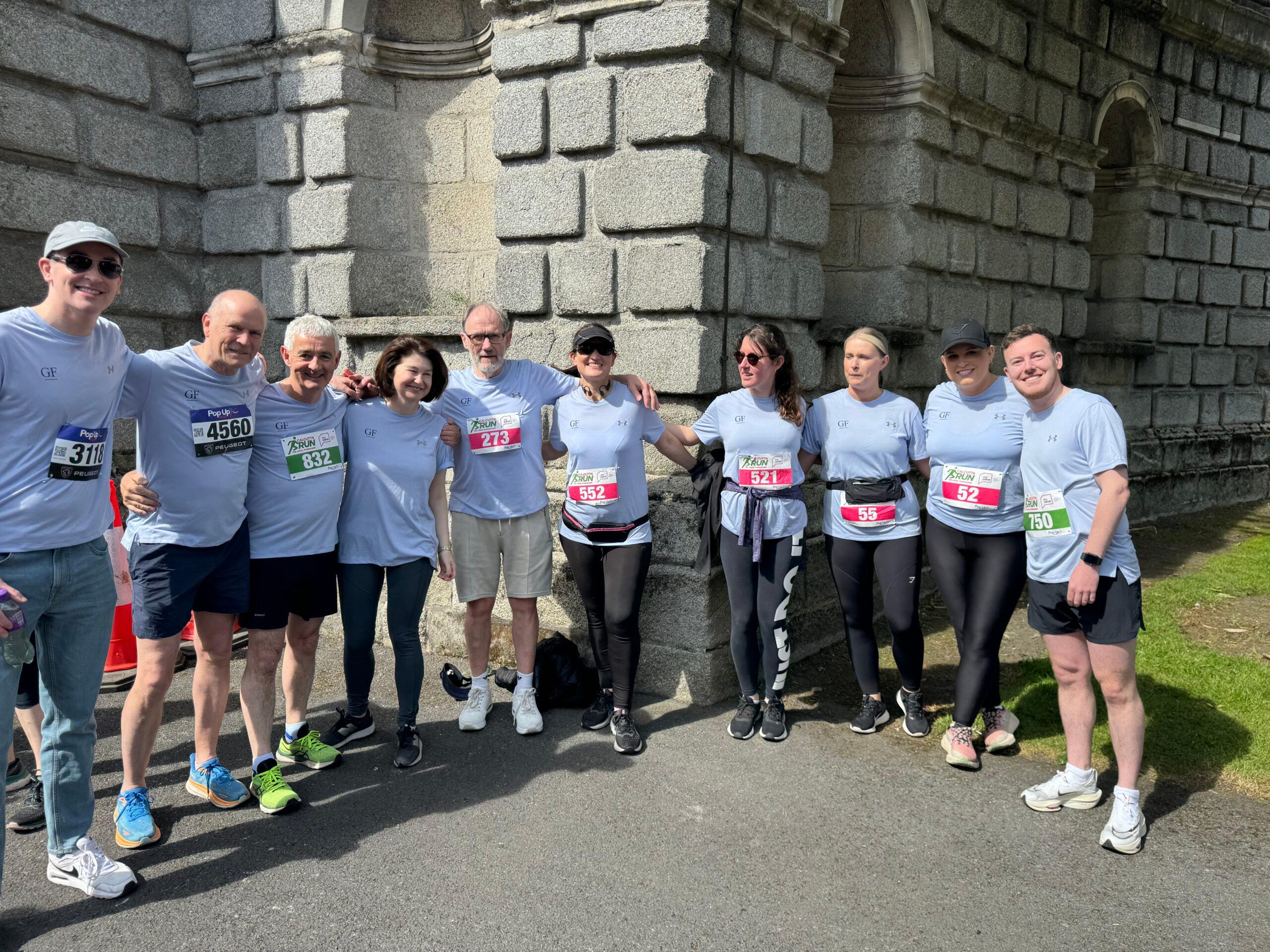 A group of ten members from the Gartlan Furey team, wearing light blue t-shirts with race numbers, posing together outdoors at the Calcutta Run 2024